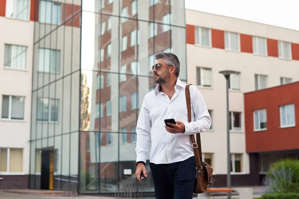 Middle-aged bearded thinking serious concentrated business man standing and smoking electronic cigarette. Blows smoke through the mouth. In-ear wireless earphone. Office centre background