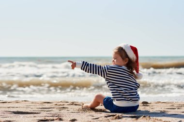Santa kırmızı şapkalı küçük kız tropik plajda eğleniyor ve sola işaret ediyor. Noel seyahat ve tatil konsepti