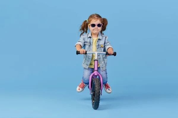 Little girl learns to keep balance while riding a bicycle. Stylish child in sunglasses  isolated on blue background