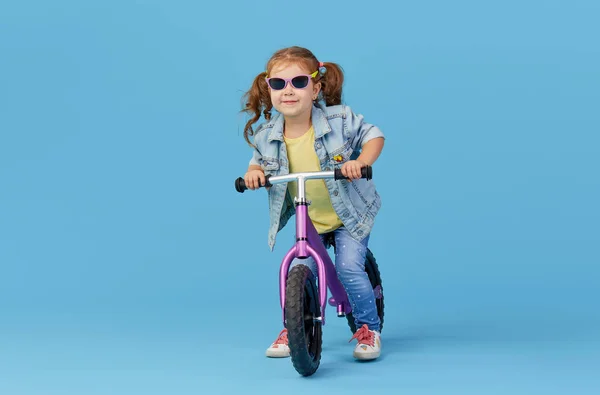 小さな女の子は自転車に乗っている間 バランスを保つことを学びます 青い背景に隔離サングラスのスタイリッシュな子供 — ストック写真