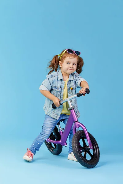 Little girl learns to keep balance while riding a bicycle. Stylish child in sunglasses  isolated on blue background
