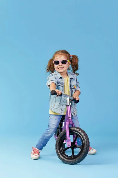 小女孩在骑自行车时学会保持平衡 时尚儿童在太阳镜隔离在蓝色背景 — 图库照片