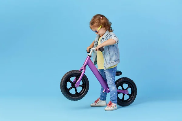 Little girl learns to keep balance while riding a bicycle. Stylish child in sunglasses  isolated on blue background