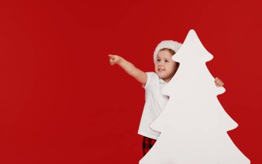 Noel Baba şapka mutlu küçük kız çocuk Noel ağaçları şeklinde boş işaret afiş arkasından gözetleme, kırmızı arka plan üzerinde izole. Metniniz için Boşluk. Sola işaret