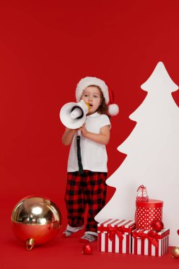 Noel Baba şapkalı mutlu çocuk kız. Noel dekorasyonunda megafonla bağıran çocuk. Mutlu xmas ve Yeni Yıl, kırmızı arka plan metin için kopya alanı ile kış tatili kavramı