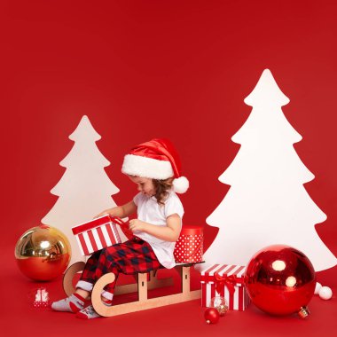 Mutlu Noeller ve Mutlu Tatiller! Sevimli küçük çocuk kız kapalı Noel ağacı dekorasyon olduğunu. Kırmızı arka planda izole edilmiş. Bir çocuk kızağa oturur ve hediye kutuları sıralar.