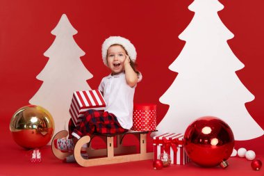 Mutlu Noeller ve Mutlu Tatiller! Sevimli küçük çocuk kız kapalı Noel ağacı dekorasyon olduğunu. Kırmızı arka planda izole edilmiş. Bir çocuk bir kızak üzerinde oturur ve hediye kutuları açılış