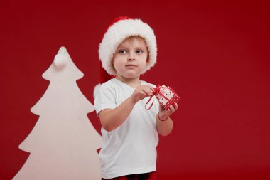 Mutlu Noeller ve Mutlu Tatiller! Sevimli küçük çocuk kapalı Noel ağacı dekorasyon olduğunu. Kırmızı arka planda izole edilmiş. Küçük Santa yardımcı bir hediye kutuları tutun