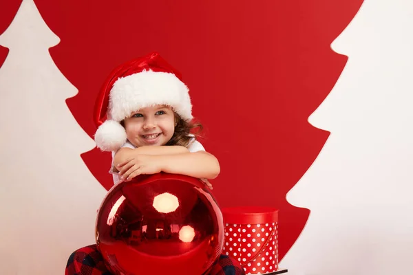Little child girl in santa claus hat sits on wooden sleigh with gift boxes and holds big christmas ball on red isolated background. blank space for text. Little Santa helper