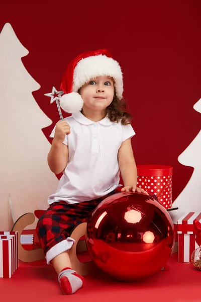 圣诞快乐 节日快乐 可爱的小女孩正在室内装饰圣诞树 在红色背景上隔离 一个孩子坐在雪橇上 打开礼物盒 — 图库照片