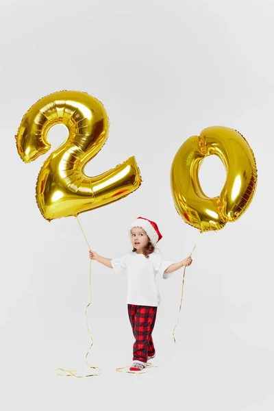 Portrait of little child girl in Santa hat posing with gold 20 twenty number balloons isolated over white background. Christmas and new year deposit installments and cashback concept. Birthday party.