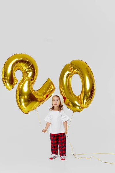 Portrait of little child girl in Santa hat posing with gold 20 twenty number balloons isolated over white background. Christmas and new year deposit installments and cashback concept. Birthday party.