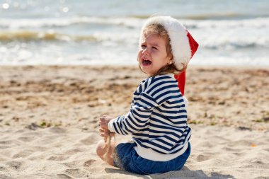 Santa şapka üzgün küçük kız okyanus ayakın tropikal bir plajda ağlıyor. Yakında bavullu bir bavul var, çocuk kayboluyor ve ailesini arıyor. Noel seyahat konsepti