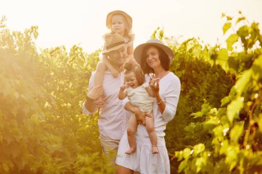 Çocuklu genç bir aile yaz günbatımı arka planında eğleniyor. Anne ve baba iki küçük kızına sarılıyor. 