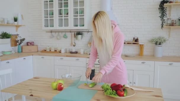 Menina loira em kigurumi quente rosa prepara salada na cozinha — Vídeo de Stock