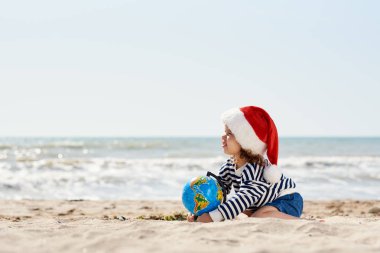 Noel Baba şapkalı mutlu küçük kız tropikal sahilde kumların üzerinde oturuyor. Çocuk Noel 'de ve yeni yılda yaz tatilinde eğleniyor. Tatil Seyahat ve macera konsepti