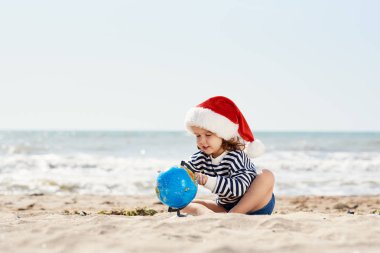 Noel Baba şapkalı mutlu küçük kız tropikal sahilde kumların üzerinde oturuyor. Çocuk Noel 'de ve yeni yılda yaz tatilinde eğleniyor. Tatil Seyahat ve macera konsepti