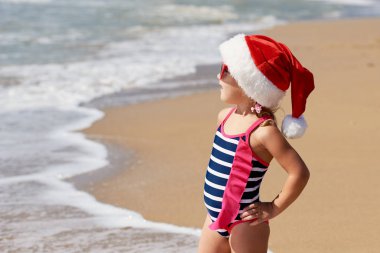 Mayo giymiş küçük bir kız çocuğu ve Noel Baba şapkası okyanusta tropik bir sahilde yürüyor. Yeni yıl ve Noel tatili kavramı. Turizm ve dinlenme