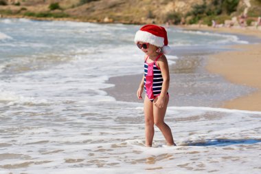 Mayo giymiş küçük bir kız çocuğu ve Noel Baba şapkası okyanusta tropik bir sahilde yürüyor. Yeni yıl ve Noel tatili kavramı. Turizm ve dinlenme