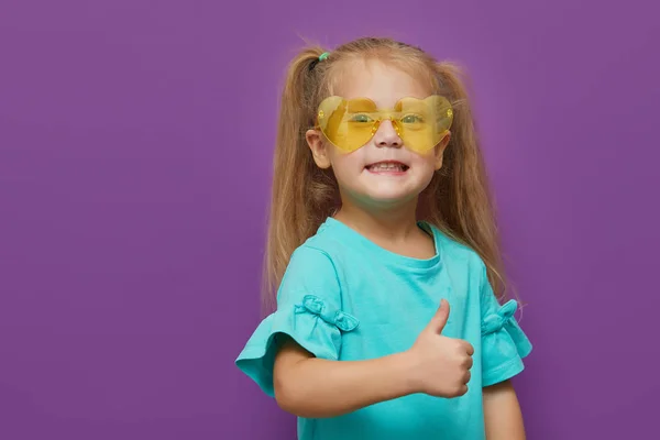 紫の背景に孤立した小さな子供の女の子の心の形のサングラスの肖像画 親指を立てて すべてはOkです 幸せな子供時代 すごいと衝撃的なコンセプト — ストック写真