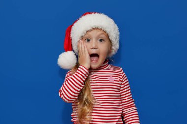 Şaşırmış ve şok olmuş, mavi arka planda tek başına duran Noel Baba şapkalı küçük kız çocuğunun portresi. Kameraya bakıyorum. Eller açık ağzın yanında. Mutlu yıllar ve Noel tatili konsepti