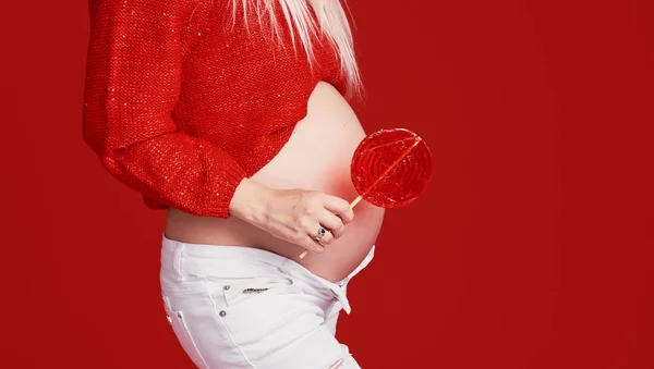 女性の健康と小児科 隔離された赤い背景に妊娠中の女性の腹のクローズアップ画像 甘いハート型のロリポップの前で開催されます クリスマスのコンセプト — ストック写真
