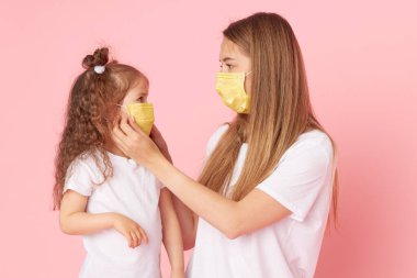 Sevdiklerinle ilgilenmek. Genç anne Covid-19 salgını sırasında koruyucu tıbbi maskeler takan küçük kıza sarılıyor. Stüdyo pembe arkaplan