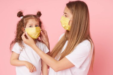 Sevdiklerinle ilgilenmek. Genç anne Covid-19 salgını sırasında koruyucu tıbbi maskeler takan küçük kıza sarılıyor. Stüdyo pembe arkaplan