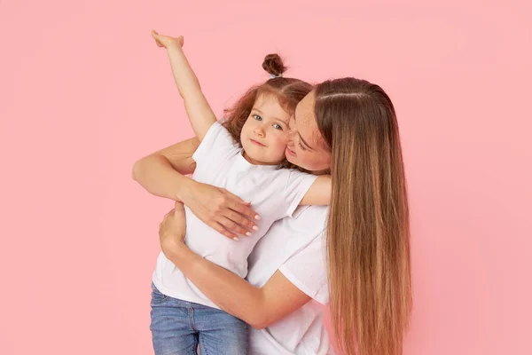 Mom and daughter in white t-shirts and jeans play and hug on a pink background. Caring for loved ones. Happy motherhood