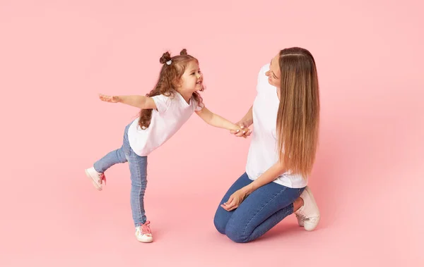 妈妈和女儿穿着白色T恤和牛仔裤在粉色背景上玩耍和拥抱 照顾所爱的人母性快乐 — 图库照片