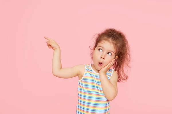 令人惊讶的可爱的小女孩的画像 背景是粉色的 看着摄像机 把手指向左边 儿童广告产品 — 图库照片