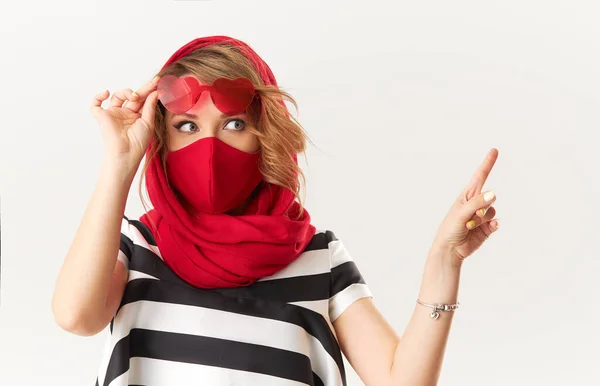 コロナウイルスの隔離中に流行のファッションの服の女性 保護スタイリッシュな赤い顔のマスクと右に指を指す白い背景にハート型のサングラスのモデル — ストック写真