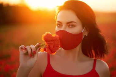 Fashion quarantine look. Young woman model in red antiviral handmade mask sniffing flowers on the background of a sunset in a field of red poppies. Summer fashion