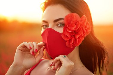 Fashion quarantine look. Young woman model in red antiviral handmade mask sniffing flowers on the background of a sunset in a field of red poppies. Summer fashion