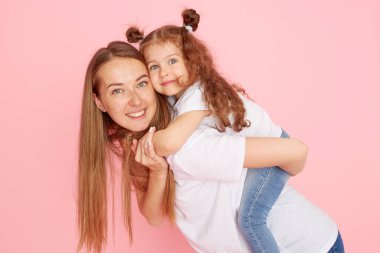 Beyaz tişörtlü ve kot pantolonlu anne ve kız pembe arka planda oynaşıp sarılıyorlar. Sevdiklerinle ilgilenmek. Mutlu annelik