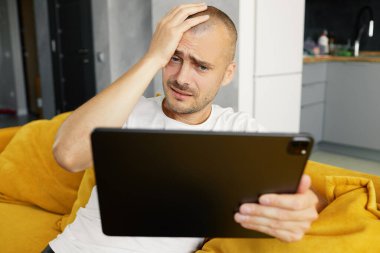 Genç üzgün adam dizüstü bilgisayarını karıştırıyor, evinde rahat bir kanepede oturuyor. Finansal değiş tokuş. Kötü haber.