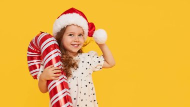 Güzel elbiseli ve Noel Baba şapkalı şirin bir kız çocuğu sarı arka planda şişme şeker kamışı şeklinde duruyor. 2021 yeni yıl tatili konsepti ve çocuklar yeni yıl hediyeleri reklamları