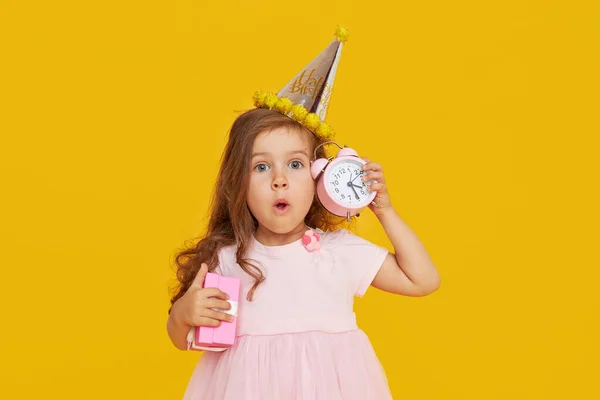 パーティーの時間だ お祝いのキャップとエレガントなドレスで楽しい女の子は彼女の誕生日を祝います 小さな目覚まし時計を保持し 黄色の背景にお祝いのパーティーを待っています — ストック写真