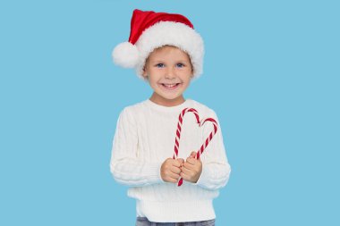 Mavi arka planda kalp şeklinde lolipop çubuğu tutan Noel Baba şapkalı sarışın bir oğlan. Çocuklarda kalp sağlığı kavramı