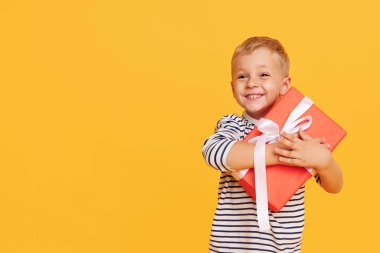 Mutlu çocuk tatilini kutlarken, heyecanlı yüzüyle hediye kutusunu açarken, sarı arka planda poz verirken.