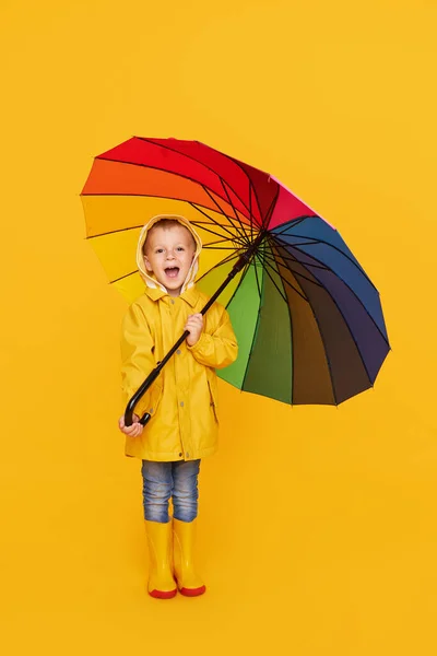 ゴムブーツの小さな幸せな感情的な陽気な子供の男の子と黄色の背景に色の傘で笑ってレインコート 梅雨の日 — ストック写真