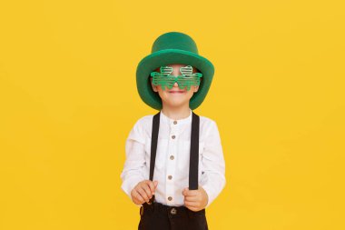 Sarı arka planda yeşil cüce şapkalı, gülümseyen küçük çocuk. Aziz Patrick Günü kutlaması. Komik surat.