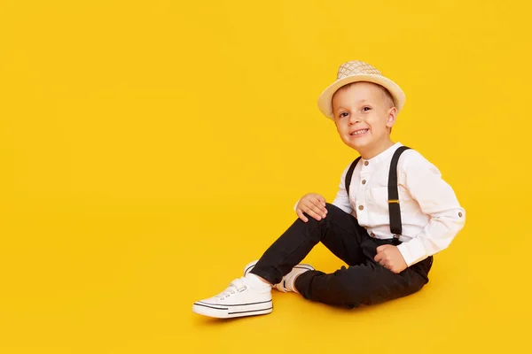 黄色の背景にポーズをとるサスペンダー 白いシャツと帽子を持つ流行の黒いズボンのスタイリッシュな子供の男の子 — ストック写真