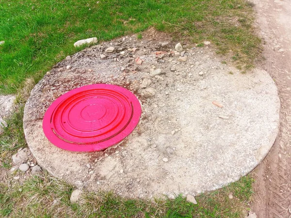 Parlak kırmızı bir rögar kapağı beton bir yapının üzerinde duruyor. Aşağıda gizli bir yangın musluğu var.
