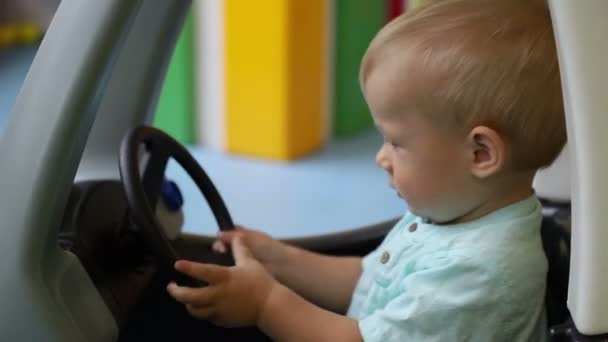 Jovem motorista. Criança fecha a porta em um carro de brinquedo e controla o volante. Menino, um ano de idade. Vista lateral. Centro de jogos infantis . — Vídeo de Stock