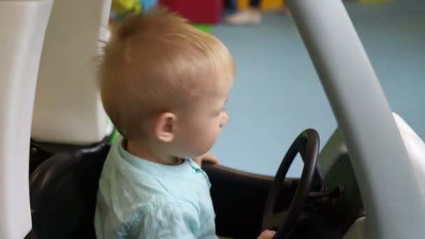 Boy sitting in a toy car. Baby closes car door and drives. Child in children’s play center. — Stock Video