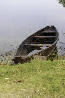 Eski, ahşap bir kayık, sakin bir gölün kayık kıyılarında huzur içinde süzülür. Teknede tek bir sarı çiçek vardır..
