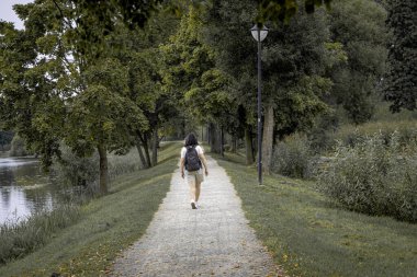 Sırt çantalı bir gezgin sakin bir günde, gür ağaçlar ve sakin bir gölle çevrili sakin bir çakıl yolunda yürüyor..
