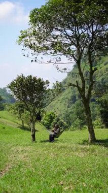 Tek başına bir erkek maceracı, etrafında yeşil çimenler ve tepeler olan sessiz bir orman bölgesinde hamakta teneke düdük adı verilen geleneksel bir müzik aleti çalar..