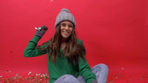 Meisje zit op de vloer op een rode achtergrond en gooit confetti in de camera — Stockvideo
